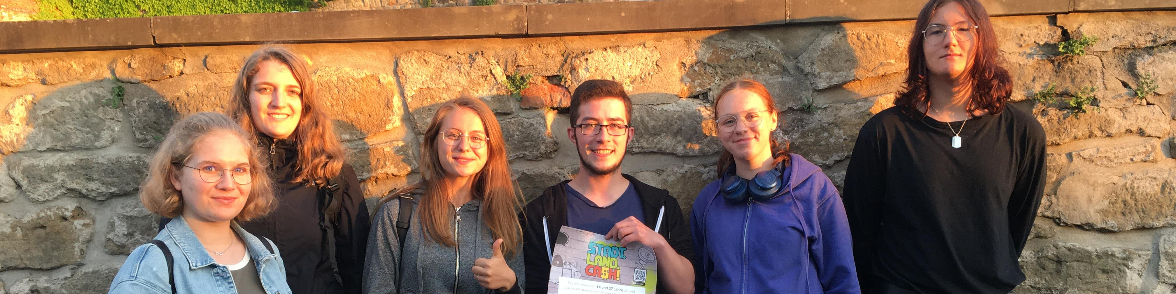 KI generiert: Das Bild zeigt eine Gruppe von sechs Personen, die vor einer alten Steinmauer stehen und in die Kamera lächeln. Eine Person in der Mitte hält ein Zertifikat oder eine Urkunde in den Händen.