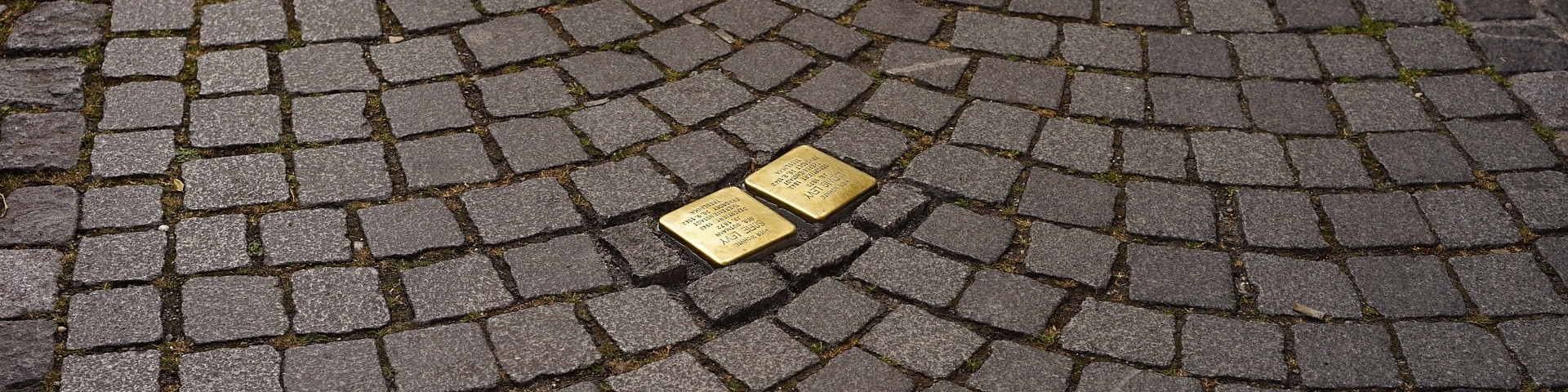 KI generiert: Das Bild zeigt zwei goldfarbene Stolpersteine, die in ein gepflastertes Gehwegfeld eingebettet sind. Diese Steine sind Teil eines Projekts zur Erinnerung an Opfer des Nationalsozialismus.