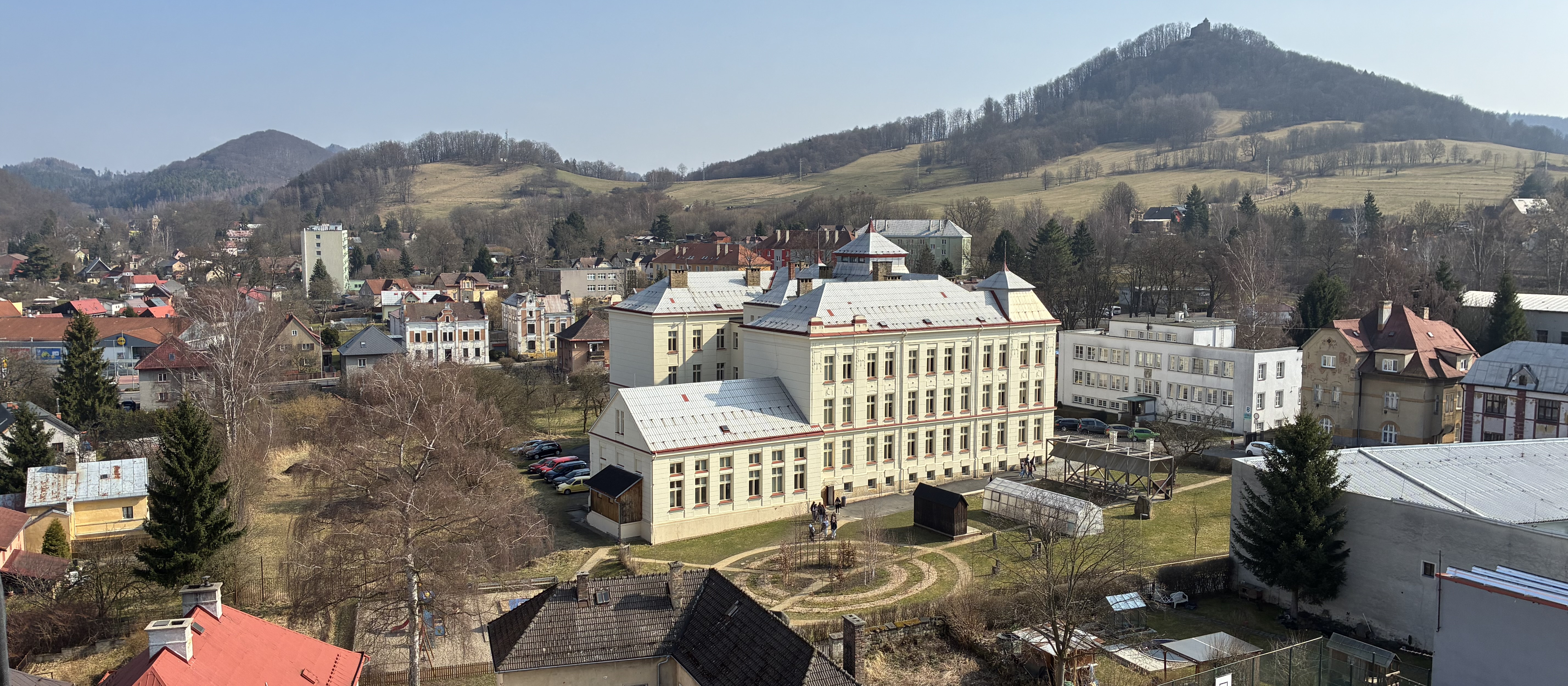 KI generiert: Das Bild zeigt eine malerische Kleinstadt mit einem großen, historischen Gebäude im Zentrum, das von einer parkähnlichen Anlage umgeben ist. Im Hintergrund sind sanfte Hügel und eine bewaldete Landschaft zu sehen.