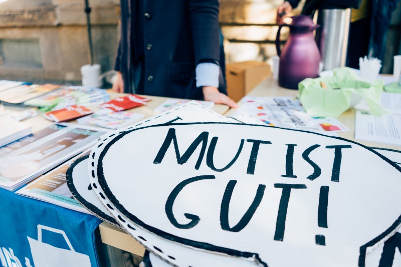 KI generiert: Das Bild zeigt einen Tisch mit verschiedenen Materialien, auf dem ein Schild mit der Aufschrift "MUT IST GUT!" im Vordergrund liegt. Im Hintergrund sind Personen zu sehen, die sich an dem Tisch befinden.