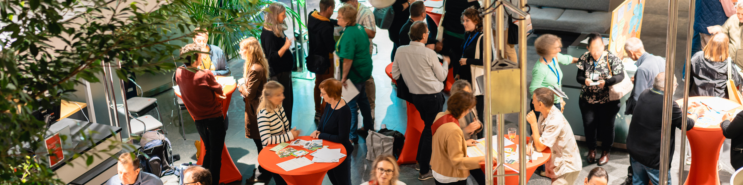 KI generiert: Das Bild zeigt eine Gruppe von Menschen, die sich in einem hellen, modernen Innenraum versammelt haben, möglicherweise bei einer Veranstaltung oder einem Treffen. Im Vordergrund sind einige Personen an Stehtischen im Gespräch, während andere in Gruppen zusammenstehen oder sitzen.