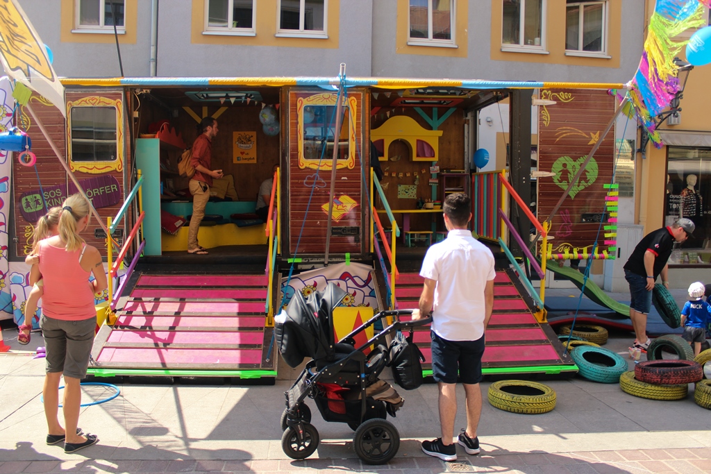 KI generiert: Das Bild zeigt eine bunte, mobile Spiel- und Kletteranlage für Kinder, die auf einem Platz vor einem Gebäude aufgestellt ist. Mehrere Erwachsene und Kinder sind in der Nähe, einige beobachten die Anlage oder bereiten sich darauf vor, sie zu nutzen.