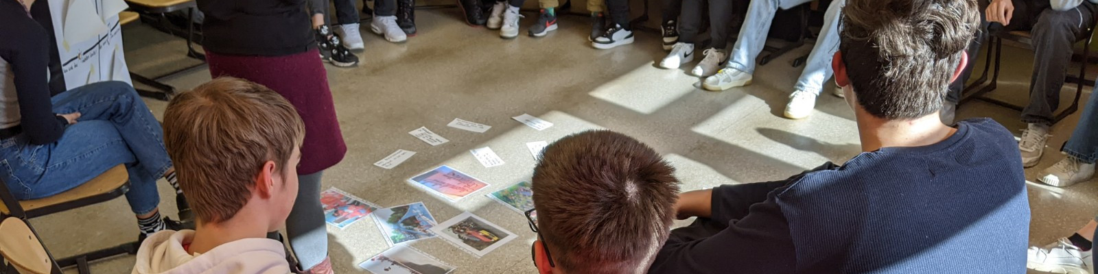 KI generiert: Das Bild zeigt eine Gruppe von Personen, die im Kreis auf dem Boden sitzen und verschiedene Bilder oder Karten betrachten. Der Hauptinhalt des Bildes ist eine interaktive Aktivität oder ein Workshop, bei dem die Teilnehmer gemeinsam etwas analysieren oder diskutieren.
