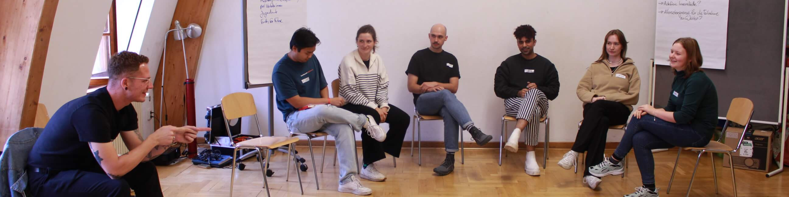 KI generiert: Das Bild zeigt eine Gruppe von sieben Personen, die in einem halbrunden Kreis auf Stühlen sitzen und sich in einem Raum mit Holzboden und großen Fenstern unterhalten. Der Hauptinhalt des Bildes ist eine Diskussion oder ein Meeting in einer informellen Atmosphäre.