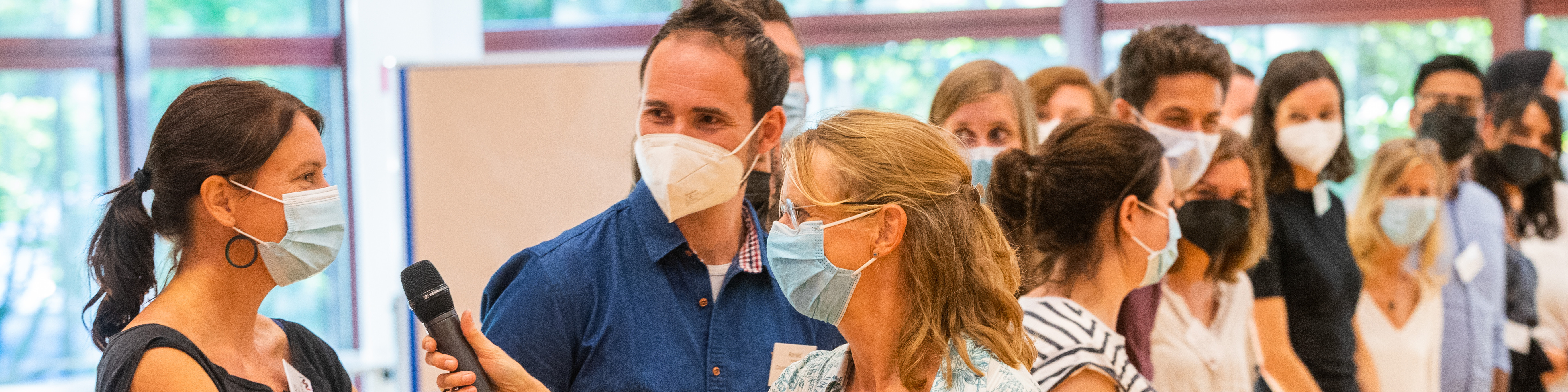 KI generiert: Das Bild zeigt eine Gruppe von Personen, die alle Gesichtsmasken tragen und in einem Innenraum stehen. Im Vordergrund halten einige Personen ein Mikrofon, was darauf hindeutet, dass sie möglicherweise an einer Veranstaltung oder Diskussion teilnehmen.