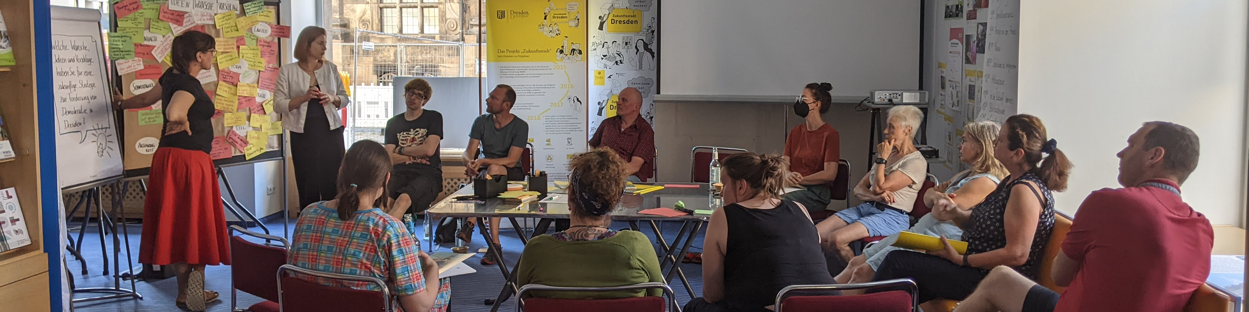 KI generiert: Das Bild zeigt eine Gruppe von Personen in einem Seminarraum, die an einer Diskussion oder einem Workshop teilnehmen. Im Vordergrund stehen zwei Frauen, eine davon präsentiert oder moderiert, während die anderen Teilnehmer an Tischen sitzen und zuhören.