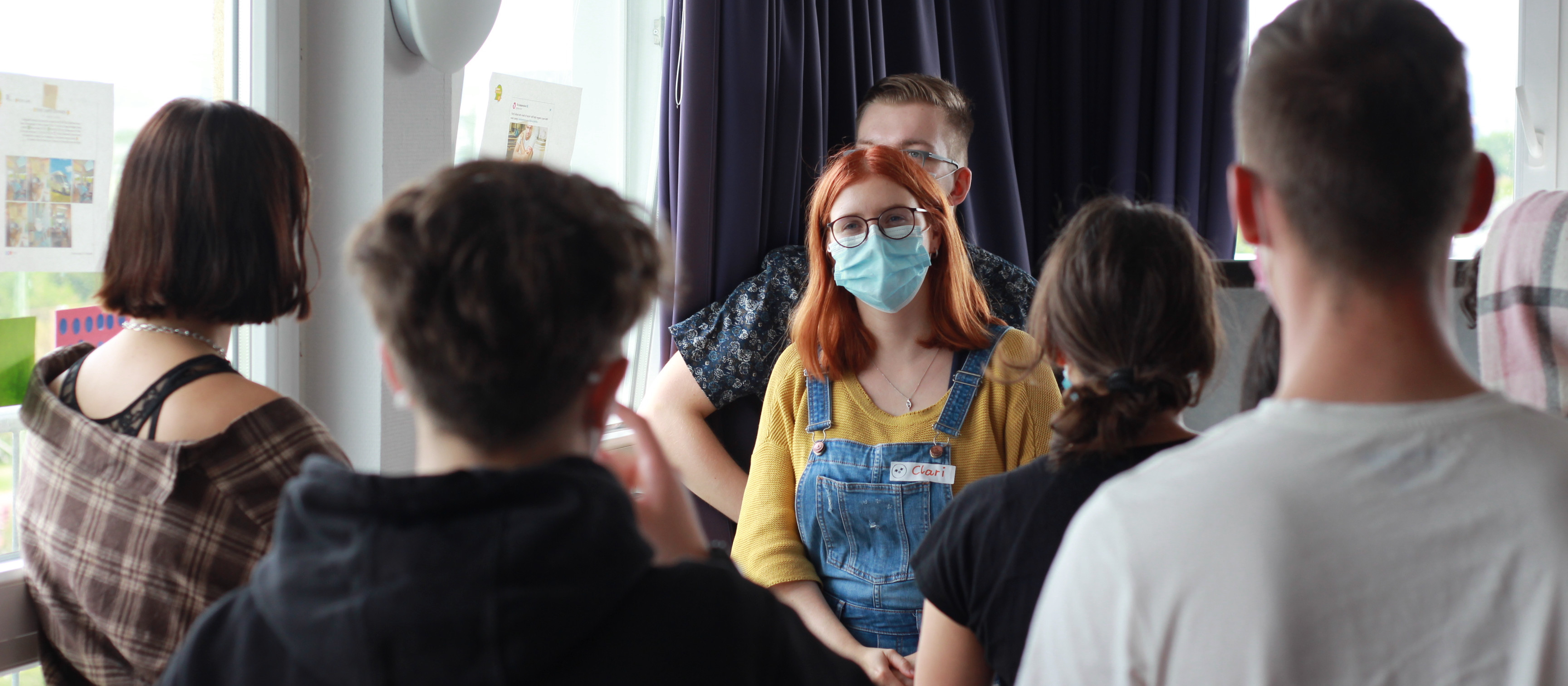 KI generiert: Das Bild zeigt eine Gruppe von Personen, die in einem Kreis sitzen und sich unterhalten, wobei eine Person mit einer Maske und Overalls im Mittelpunkt steht. Der Hauptinhalt des Bildes ist eine Diskussion oder ein Treffen in einer informellen Umgebung.