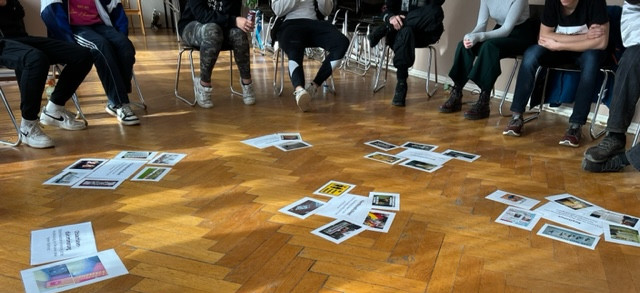 KI generiert: Das Bild zeigt eine Gruppe von Personen, die im Kreis auf Stühlen sitzen und auf den Boden schauen, wo zahlreiche Fotos und Dokumente verstreut liegen. Der Hauptinhalt des Bildes scheint eine Gruppenaktivität oder ein Workshop zu sein, bei dem die Teilnehmer die Materialien auf dem Boden betrachten und diskutieren.