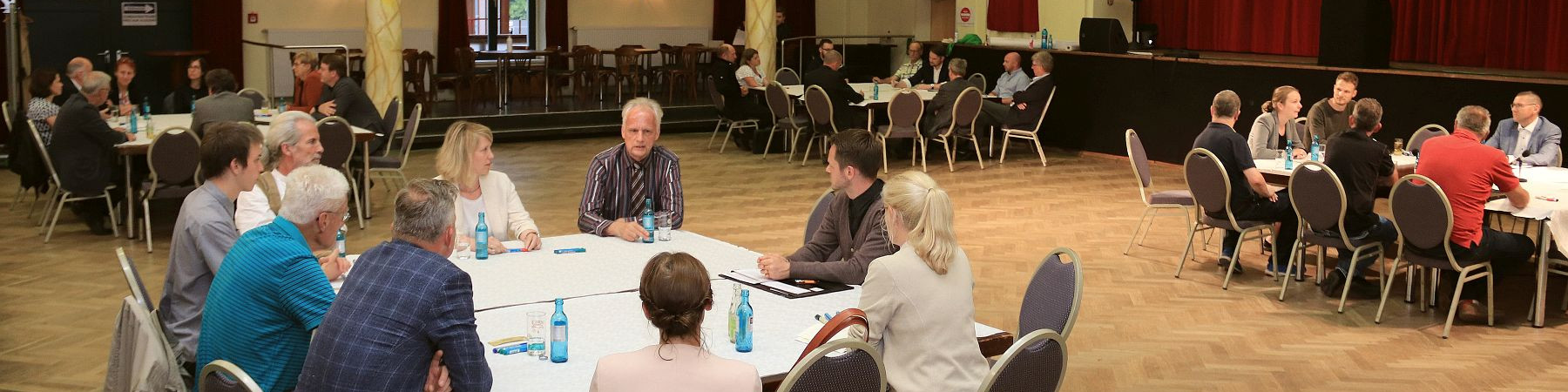KI generiert: Das Bild zeigt eine Gruppe von Personen, die in einem großen Raum an mehreren runden Tischen sitzen und sich unterhalten. Der Hauptinhalt des Bildes ist eine Versammlung oder ein Treffen, bei dem die Teilnehmer in kleinen Gruppen diskutieren.
