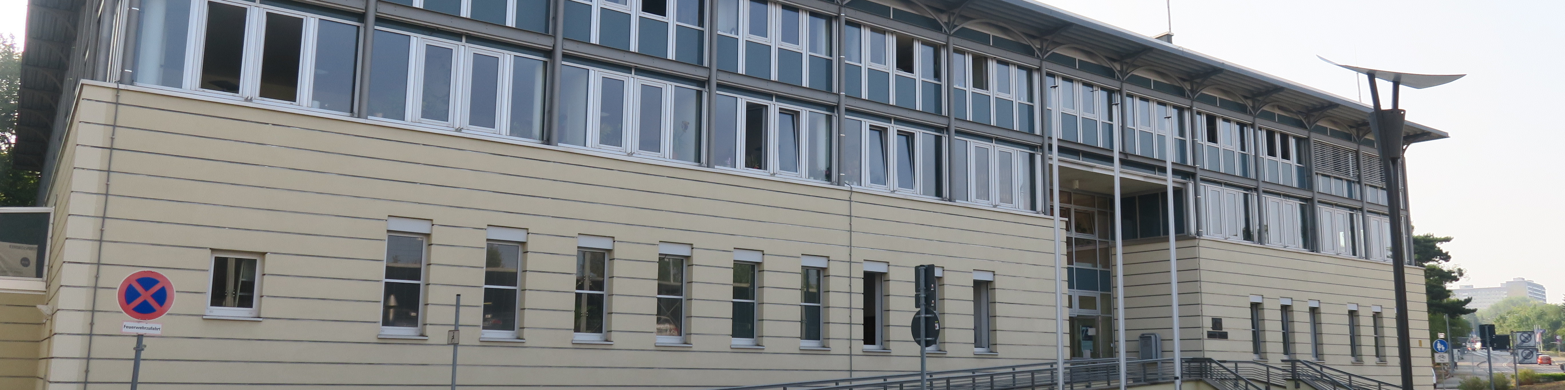 KI generiert: Das Bild zeigt ein modernes, mehrstöckiges Gebäude mit vielen Fenstern und einer Fassade aus hellen Paneelen. Im Vordergrund ist ein Verkehrsschild zu sehen, das auf ein Parkverbot hinweist. Der Hauptinhalt des Bildes ist das Gebäude.