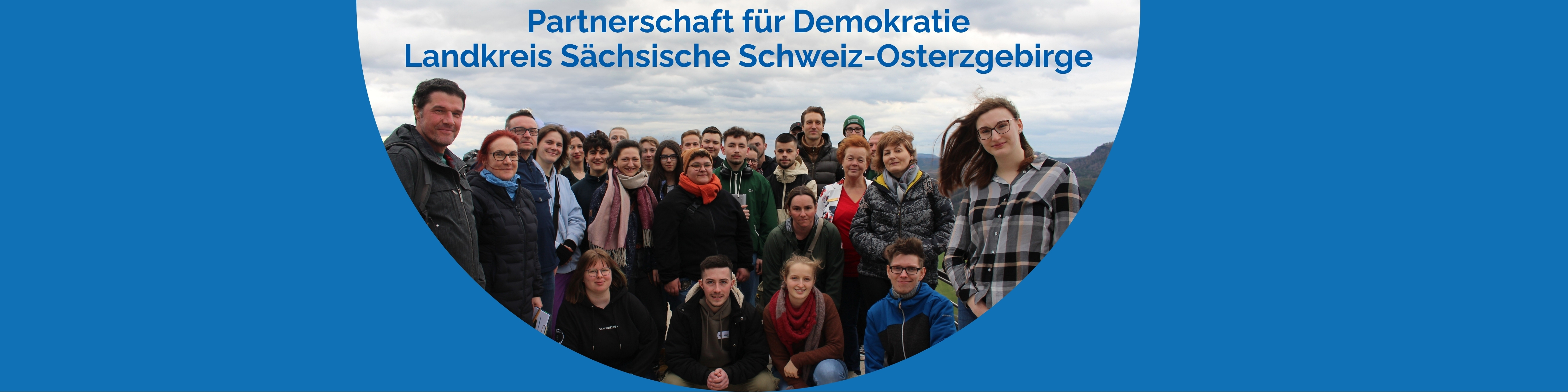 KI generiert: Das Bild zeigt eine Gruppe von Menschen, die gemeinsam für ein Foto posieren, mit einem Text darüber, der "Partnerschaft für Demokratie Landkreis Sächsische Schweiz-Osterzgebirge" lautet. Der Hauptinhalt des Bildes ist die Darstellung einer Gruppe, die sich vermutlich für demokratische Werte und Zusammenarbeit im genannten Landkreis einsetzt.