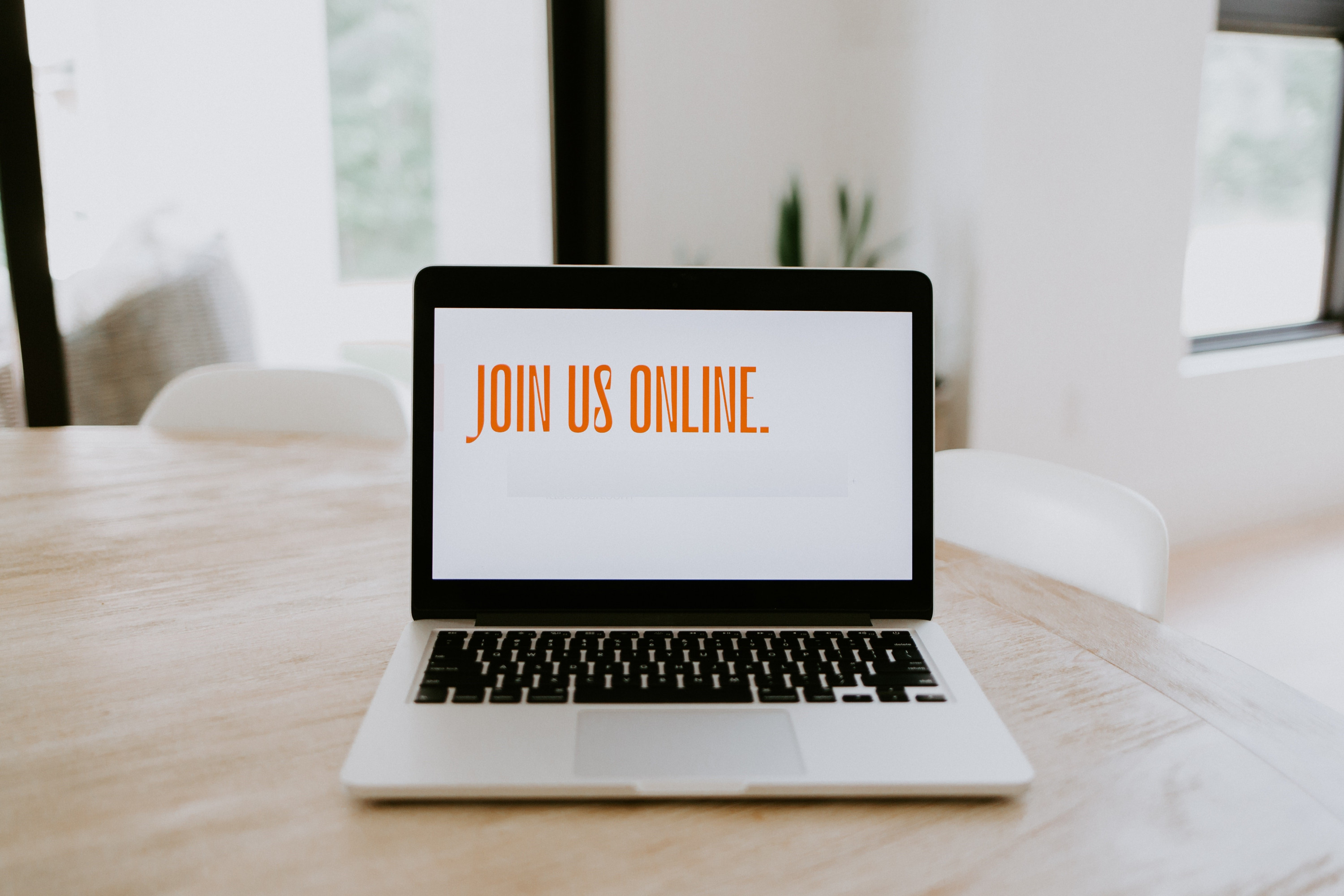 KI generiert: Das Bild zeigt einen Laptop auf einem Holztisch mit der Bildschirmanzeige "JOIN US ONLINE." Im Hintergrund sind ein Stuhl und ein Fenster zu sehen. Der Hauptinhalt des Bildes ist die Einladung, online teilzunehmen.