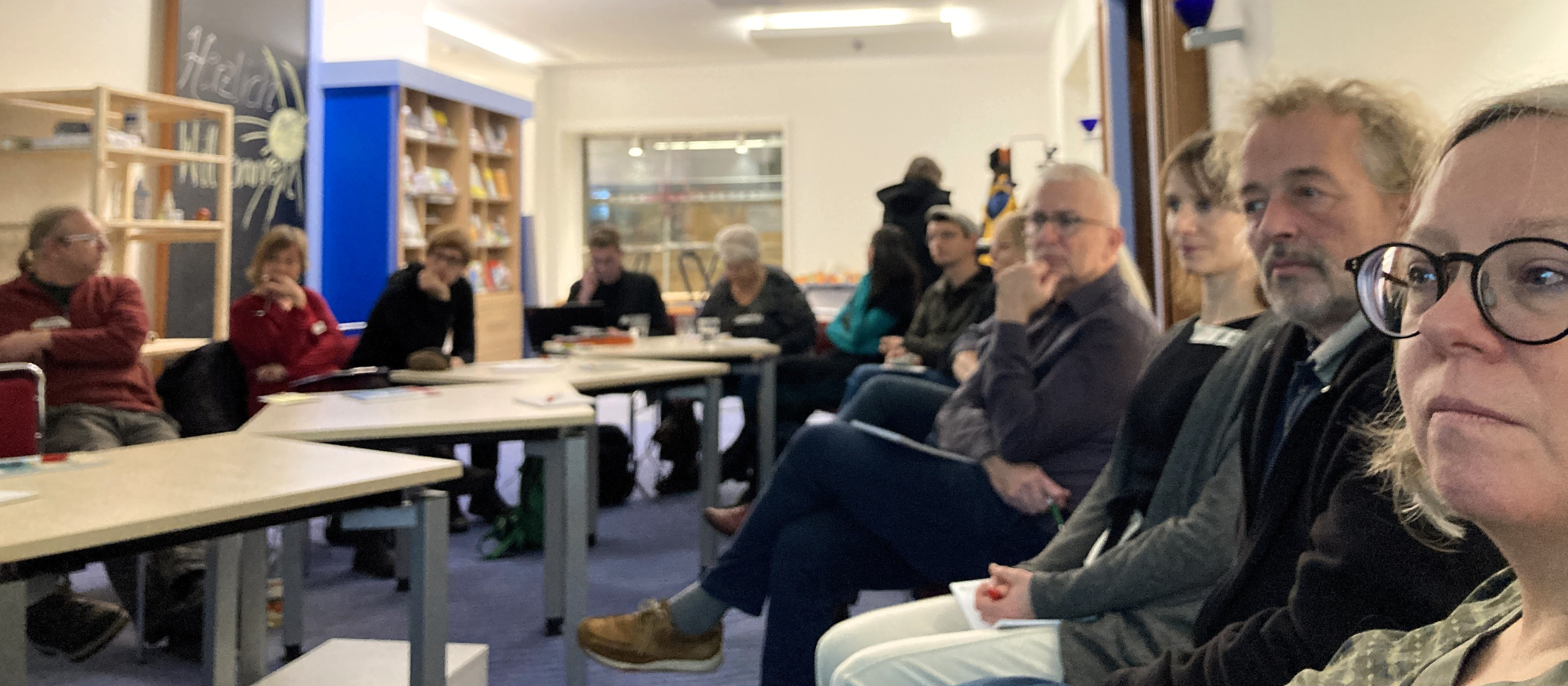 KI generiert: Das Bild zeigt eine Gruppe von Personen, die in einem Raum mit Tischen und Stühlen sitzen, möglicherweise bei einer Besprechung oder einem Workshop. Der Hauptinhalt des Bildes ist eine Versammlung oder ein Treffen in einem Innenraum.