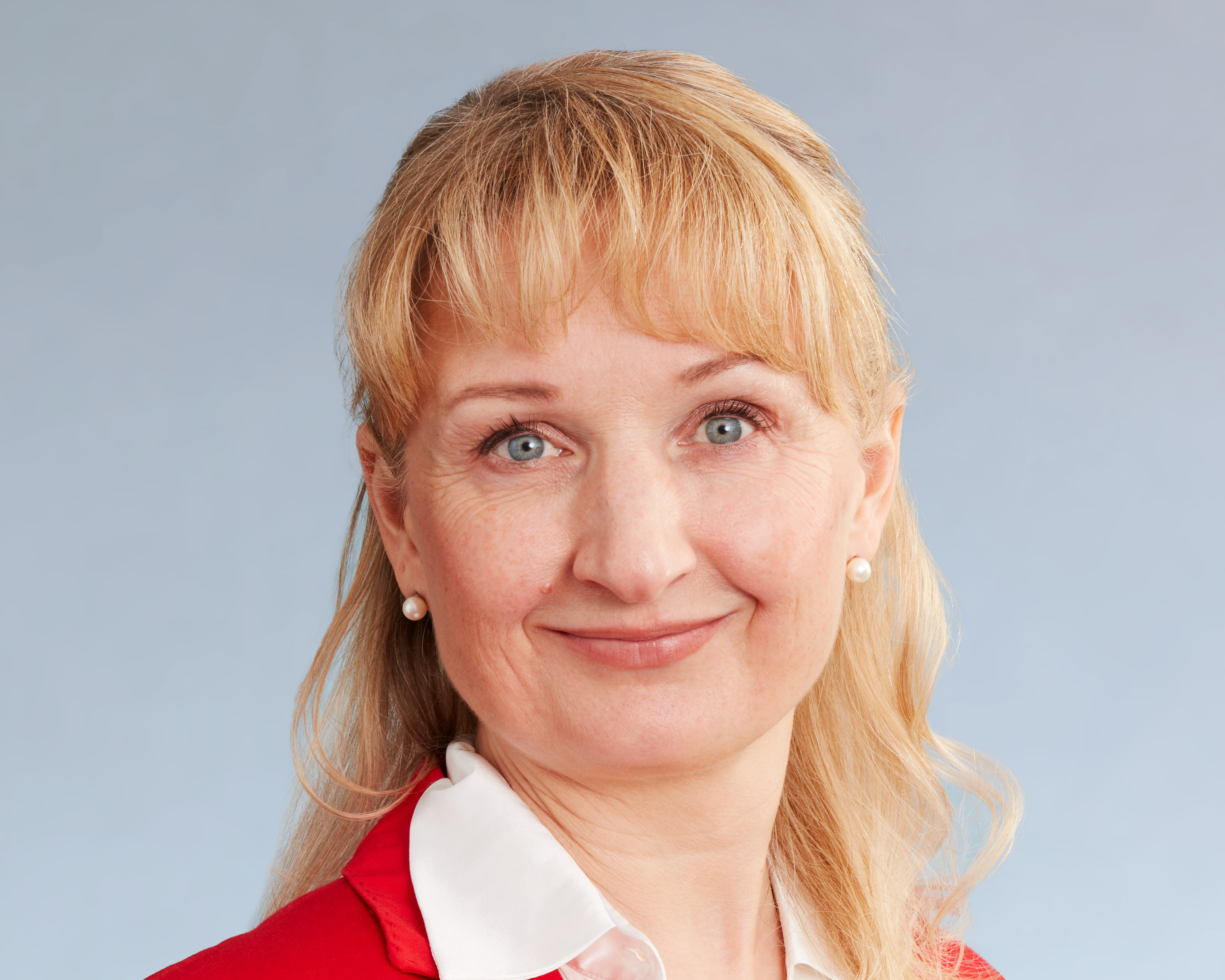 KI generiert: Das Bild zeigt eine lächelnde Frau mit blonden Haaren, die einen roten Blazer über einem weißen Hemd trägt. Der Hauptinhalt des Bildes ist das Porträt dieser Frau vor einem neutralen, hellblauen Hintergrund.