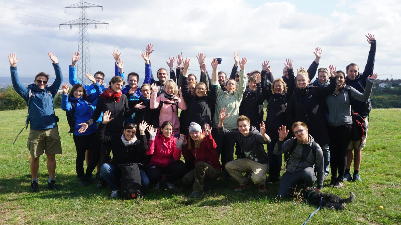 KI generiert: Das Bild zeigt eine Gruppe von Menschen, die draußen auf einer Wiese stehen und sich freudig mit hochgereckten Armen präsentieren. Im Vordergrund ist ein Hund zu sehen, und im Hintergrund sind Strommasten und eine leicht bewölkte Himmel zu erkennen.