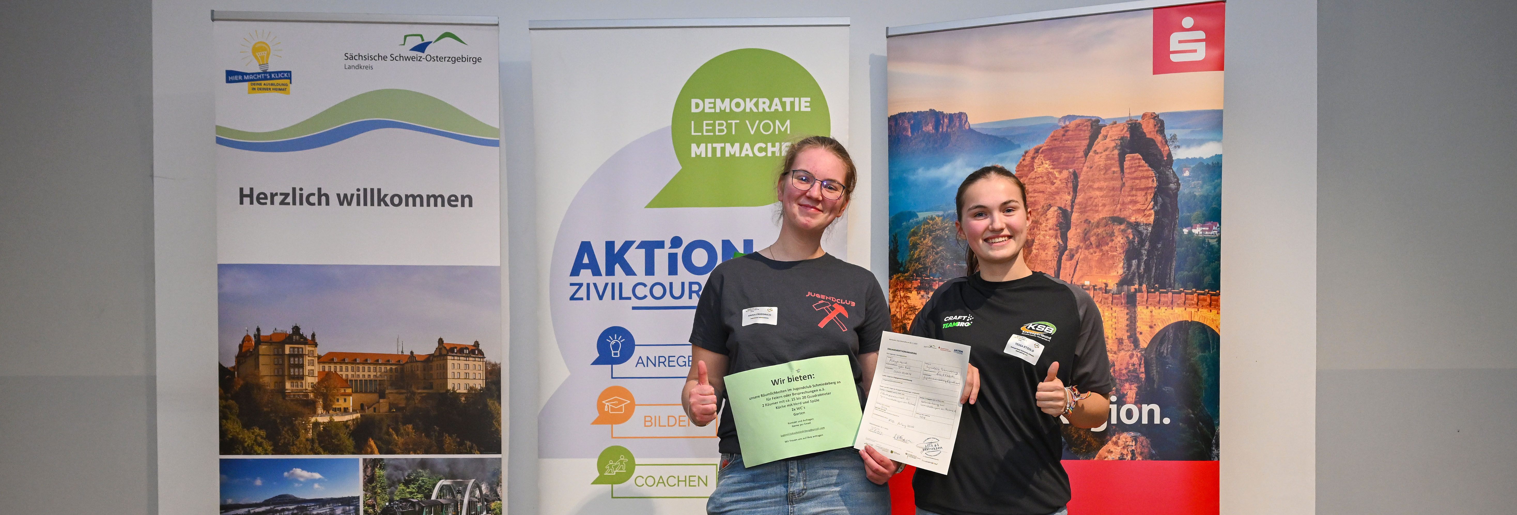 KI generiert: Das Bild zeigt zwei Personen, die lächelnd vor mehreren Roll-up-Bannern stehen und jeweils ein Dokument in der Hand halten. Die Banner beinhalten Begrüßungen und Informationen zu verschiedenen Themen wie Demokratie, Zivilcourage und regionalen Angeboten.