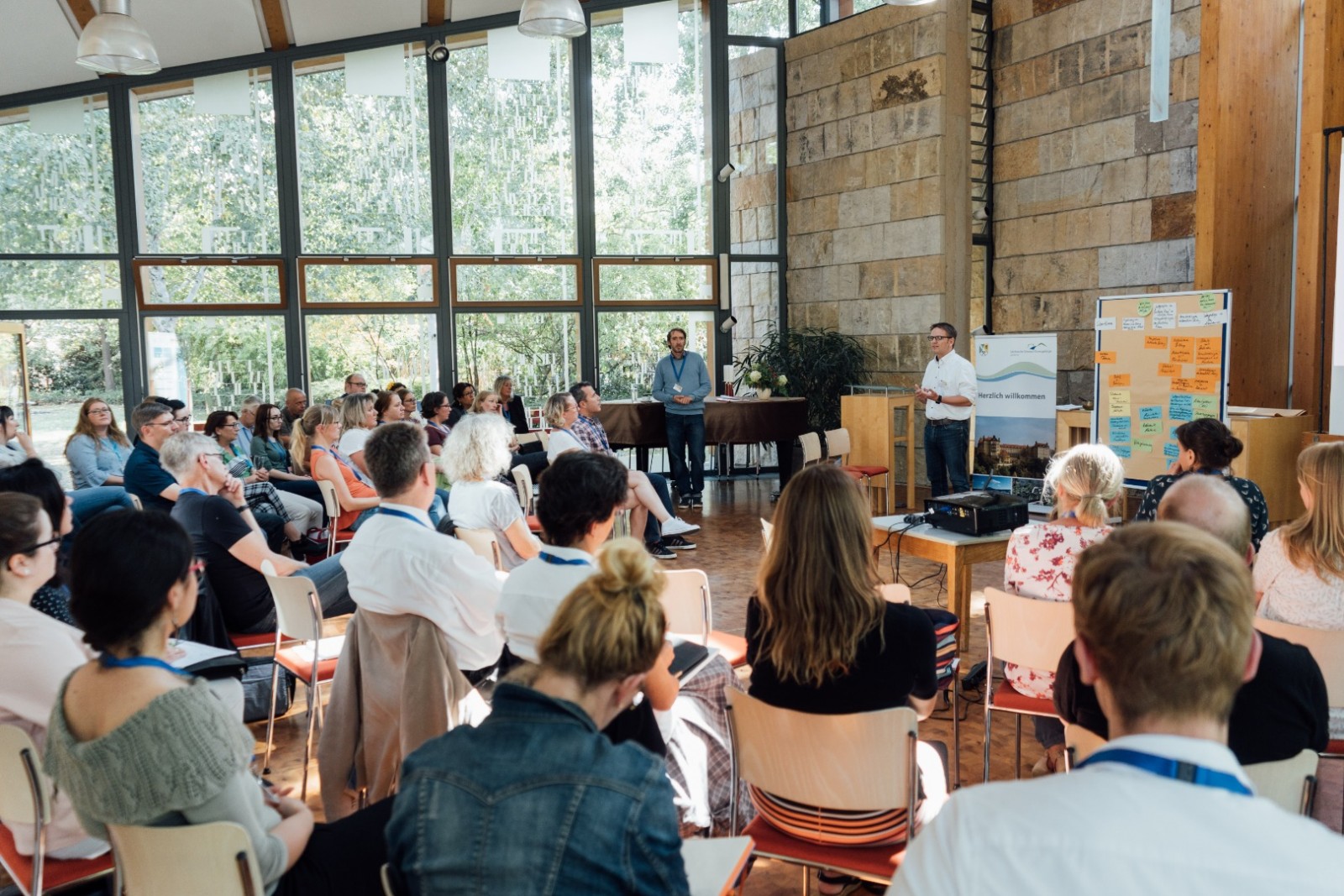 KI generiert: Das Bild zeigt eine Gruppe von Menschen, die in einem hellen, modernen Raum mit großen Fenstern an einer Präsentation oder einem Seminar teilnehmen. Der Hauptinhalt ist eine Versammlung oder ein Workshop, bei dem ein Mann vorne steht und zu den Anwesenden spricht.