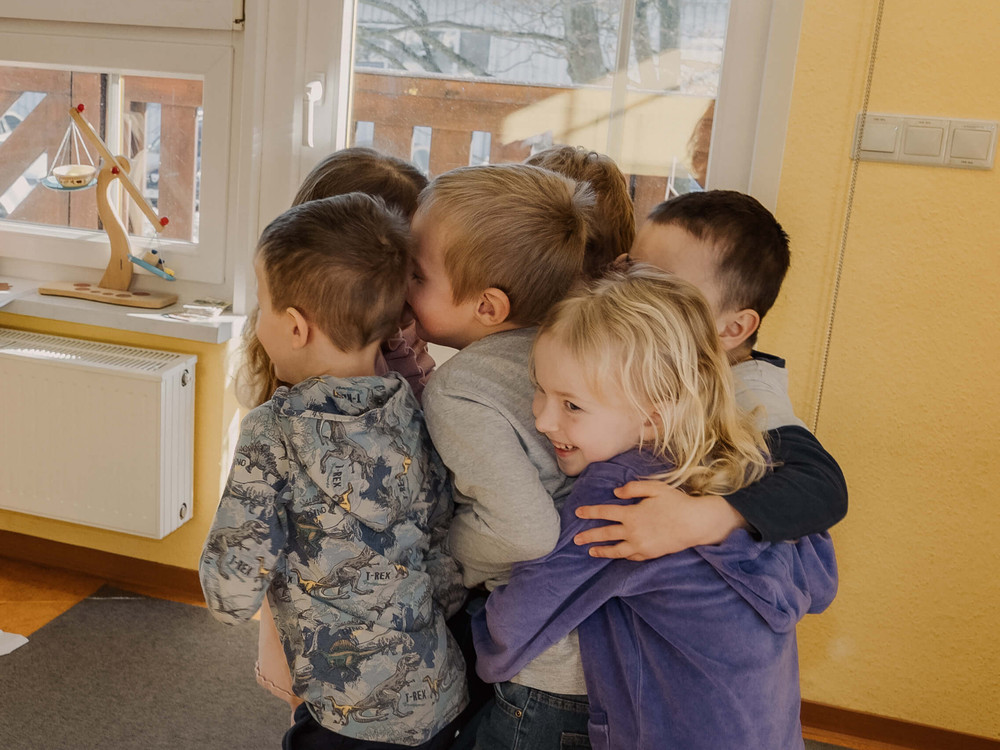 KI generiert: Das Bild zeigt eine Gruppe von Kindern, die sich in einem Raum umarmen und gemeinsam lachen. Im Hintergrund sind ein Fenster und ein Regal mit einem Spielzeug zu sehen.