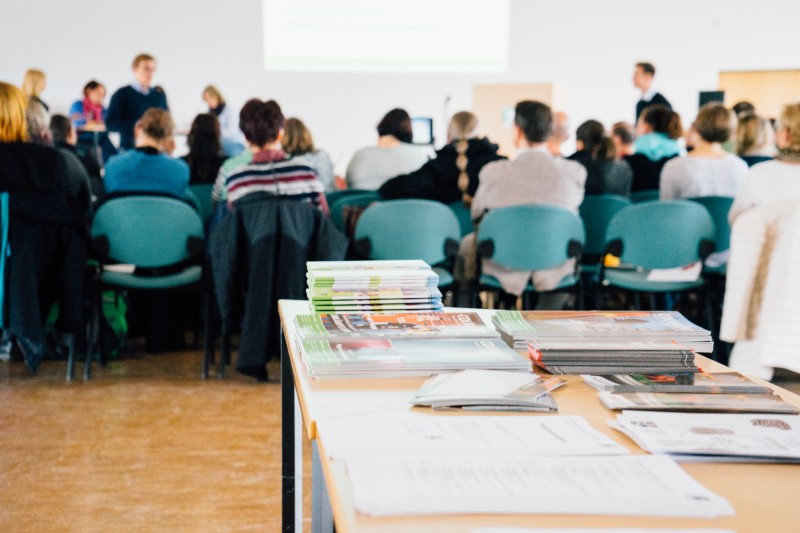KI generiert: Das Bild zeigt eine Gruppe von Personen, die in einem Seminarraum oder einer Konferenz sitzen und einer Präsentation oder Diskussion folgen. Im Vordergrund liegen mehrere Stapel mit Dokumenten oder Broschüren auf einem Tisch.