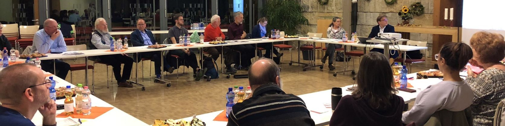 KI generiert: Das Bild zeigt eine Gruppe von Personen, die an langen Tischen in einem Raum mit großen Fenstern sitzen und sich in einer Besprechung oder einem Meeting befinden. Der Hauptinhalt des Bildes ist eine formelle Zusammenkunft oder Konferenz.