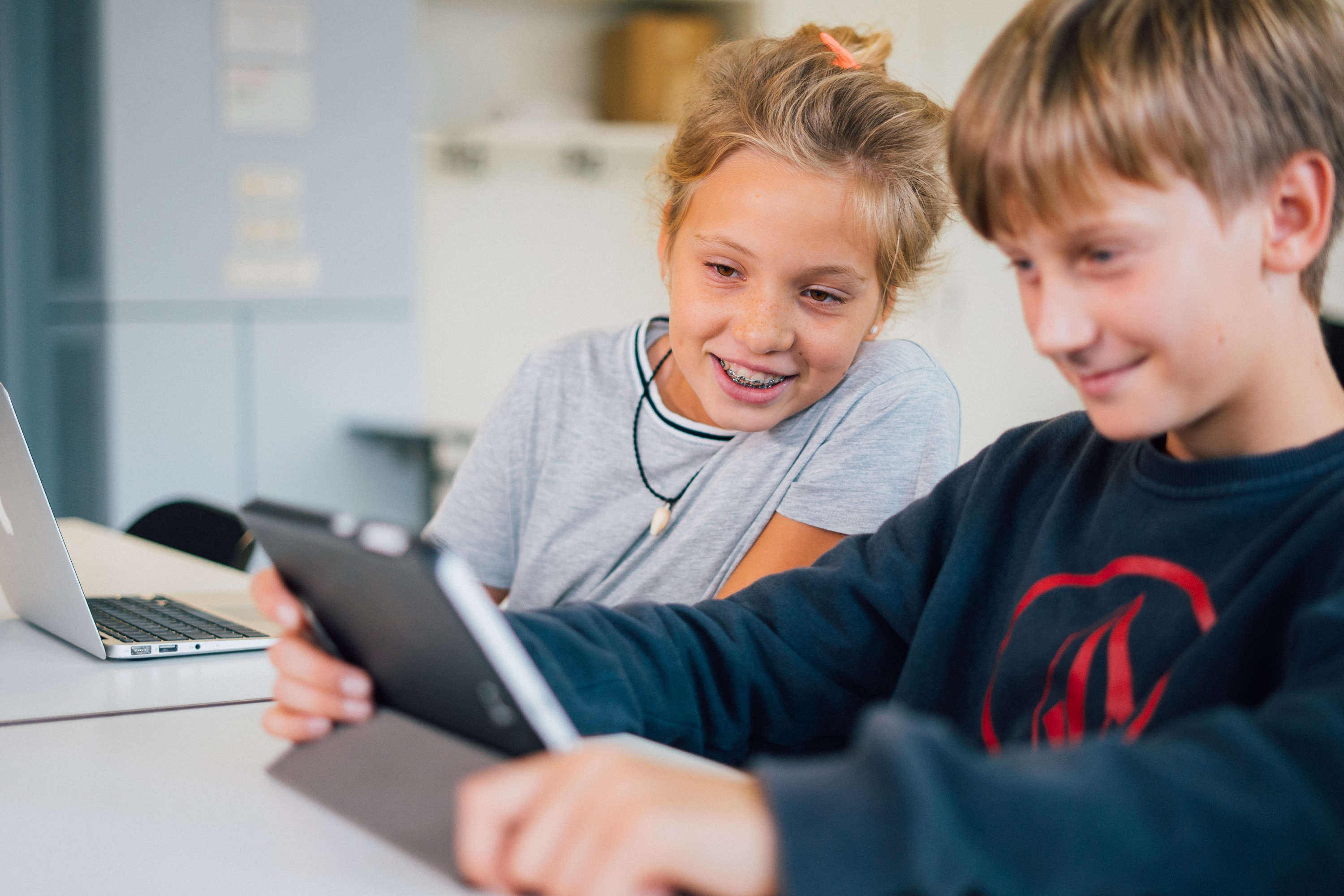 KI generiert: Das Bild zeigt zwei Kinder, die gemeinsam an einem Tisch sitzen und sich auf einen Tablet-PC konzentrieren, während ein Laptop in der Nähe liegt. Der Hauptinhalt des Bildes ist die Interaktion der Kinder mit dem Tablet, was auf eine Lern- oder Spielsituation hindeuten könnte.