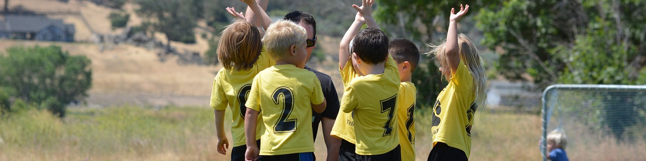 KI generiert: Das Bild zeigt eine Gruppe von Kindern in gelben Sporttrikots, die auf einem Feld stehen und sich mit hochgereckten Armen freuen. Im Hintergrund sind Bäume, ein Haus und ein Tor zu erkennen.