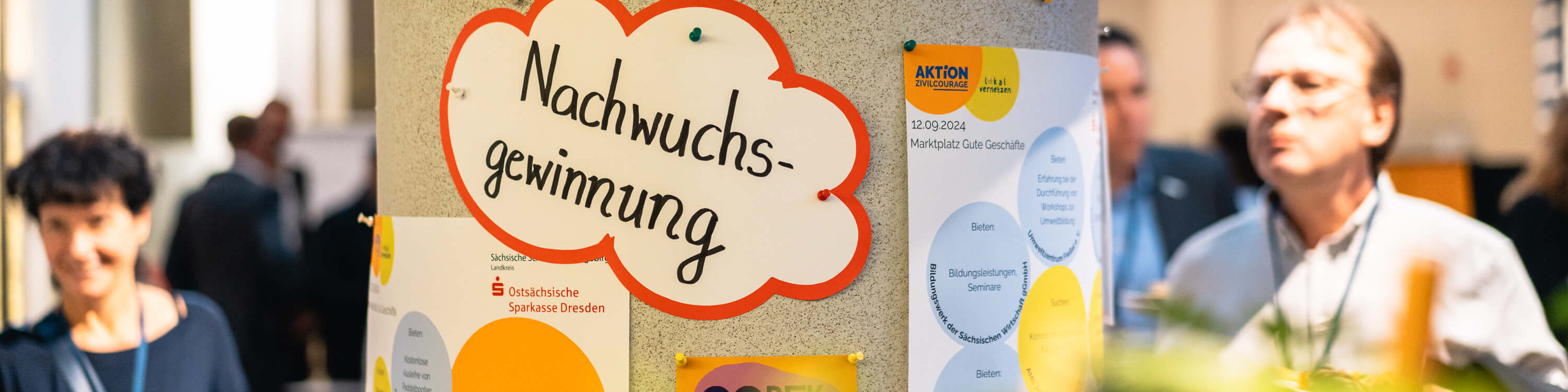 KI generiert: Das Bild zeigt eine Gruppe von Personen auf einer Veranstaltung, bei der ein Plakat mit der Aufschrift "Nachwuchsgewinnung" (Gewinnung von Nachwuchs) im Vordergrund hängt. Im Hintergrund sind weitere Plakate und Menschen zu sehen, die sich unterhalten oder Informationen betrachten.