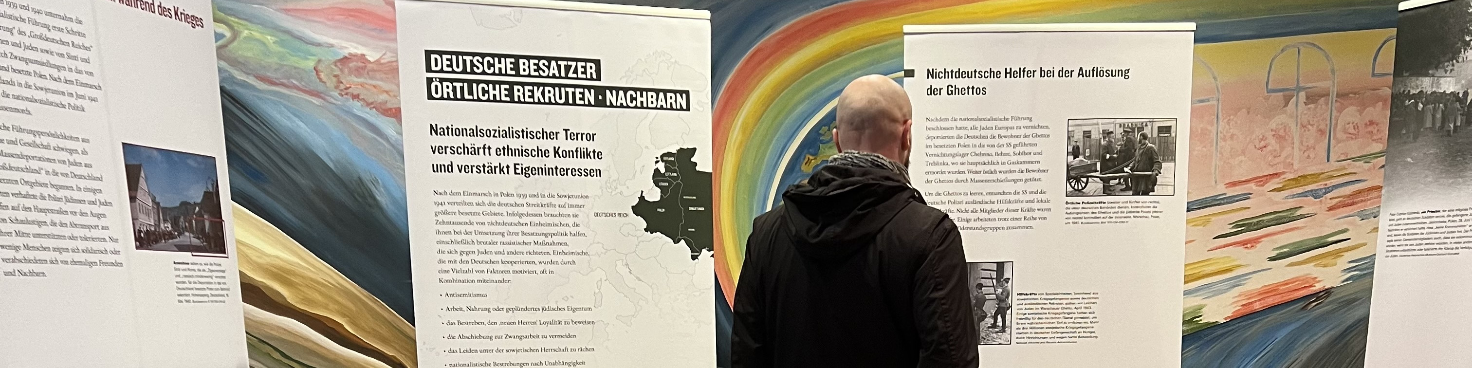 KI generiert: Das Bild zeigt einen Mann, der eine Ausstellung besucht und sich Informationsplakate ansieht, die sich auf die deutsche Besatzung und lokale Rekrutierung beziehen. Der Hauptinhalt der Ausstellung scheint historische Konflikte und Ereignisse während der deutschen Besatzung zu thematisieren.