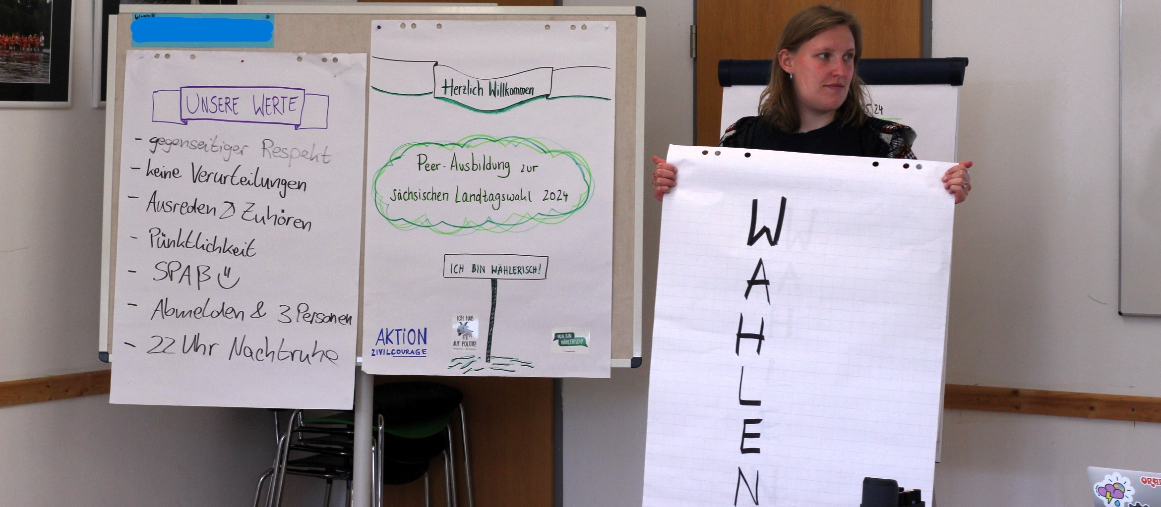 KI generiert: Das Bild zeigt eine Person, die ein Flipchart mit dem Wort "WAHLEN" hochhält, während im Hintergrund weitere Flipcharts mit Notizen und Plakaten zu sehen sind, die sich auf Werte und eine Wahl im Jahr 2024 beziehen. Der Hauptinhalt des Bildes ist die Betonung von Werten und die Aufforderung zur Wahlbeteiligung.