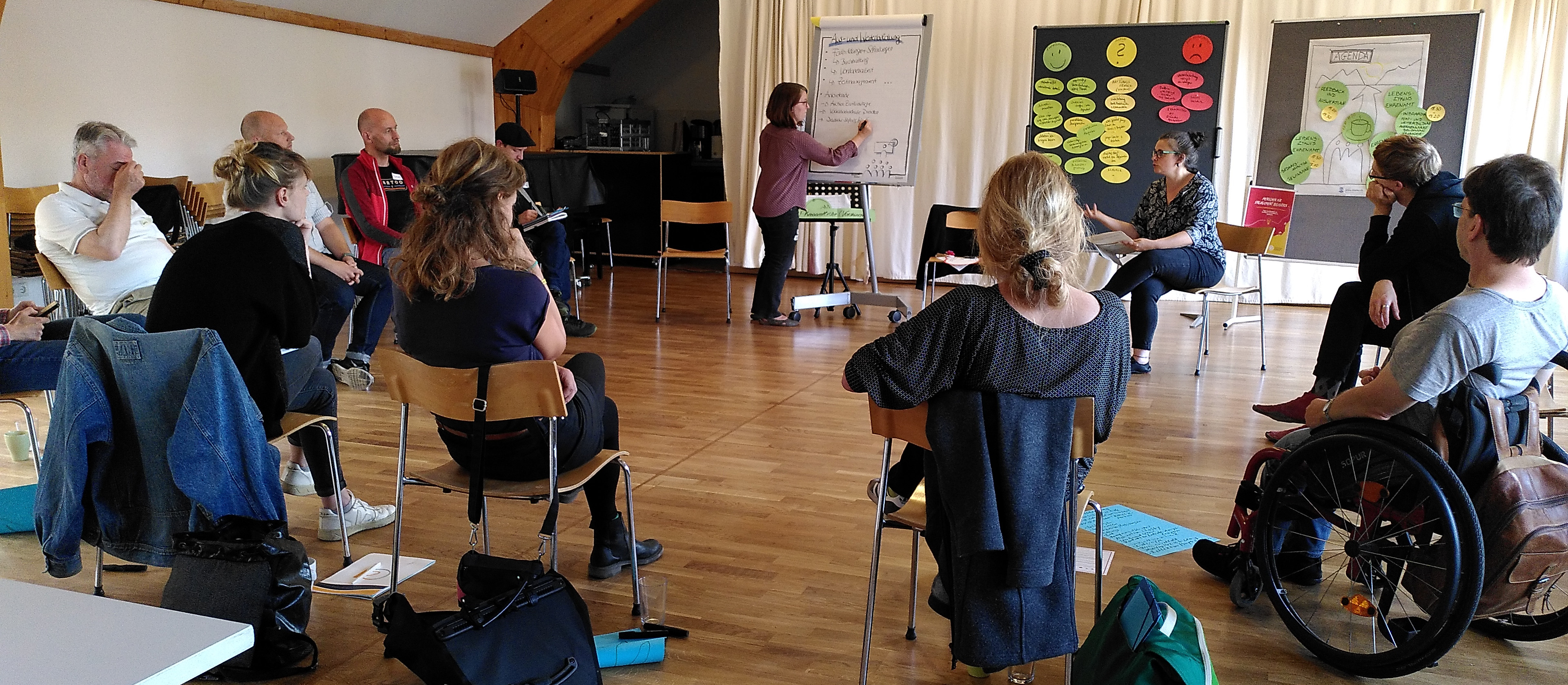 KI generiert: Das Bild zeigt eine Gruppe von Menschen in einem Seminarraum, die an einer Präsentation oder einem Workshop teilnehmen. Eine Person steht vorne und präsentiert Inhalte an einem Flipchart, während die anderen Teilnehmer aufmerksam zuhören und sich Notizen machen.