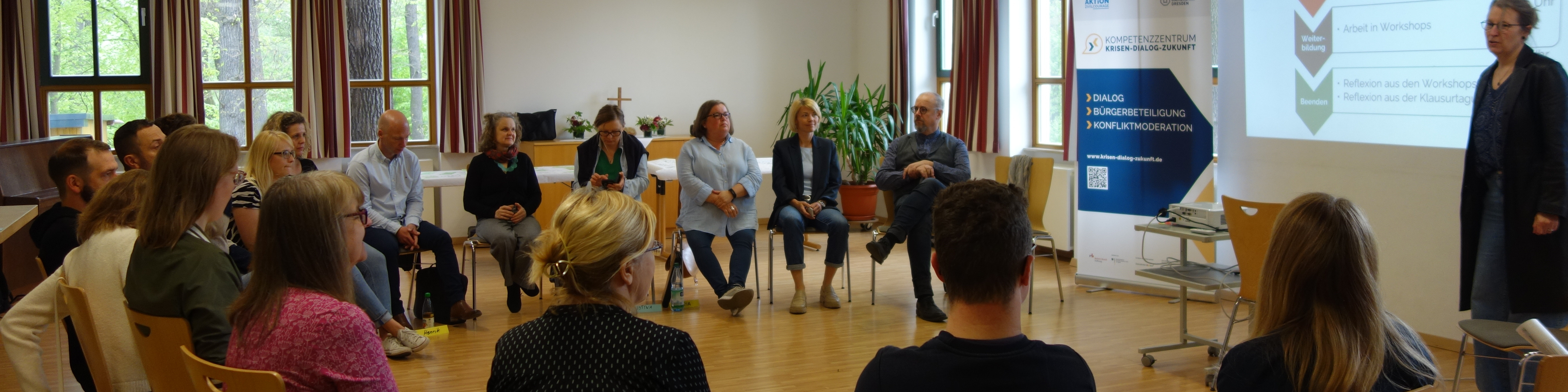 KI generiert: Das Bild zeigt eine Gruppe von Menschen in einem Seminarraum, wobei einige auf Stühlen im Kreis sitzen und eine Person vorne steht und präsentiert. Der Hauptinhalt des Bildes ist eine Diskussion oder ein Workshop in einer Bildungs- oder Schulungseinrichtung.