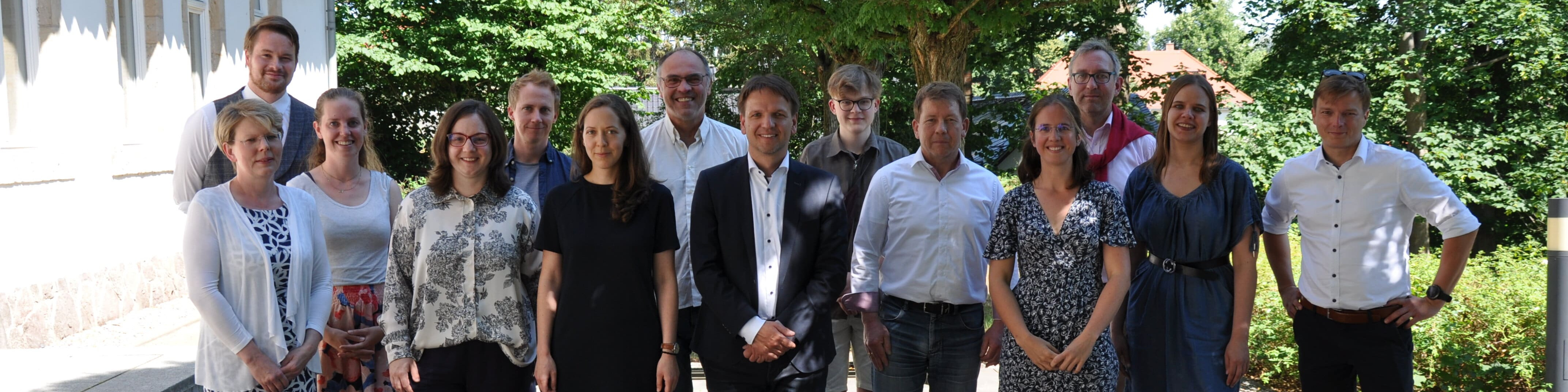 KI generiert: Das Bild zeigt eine Gruppe von 14 Personen, die in einer Reihe vor einem Gebäude und grünen Bäumen stehen und in die Kamera lächeln. Der Hauptinhalt des Bildes ist eine Gruppenfotografie, die vermutlich bei einer offiziellen oder geselligen Veranstaltung entstanden ist.