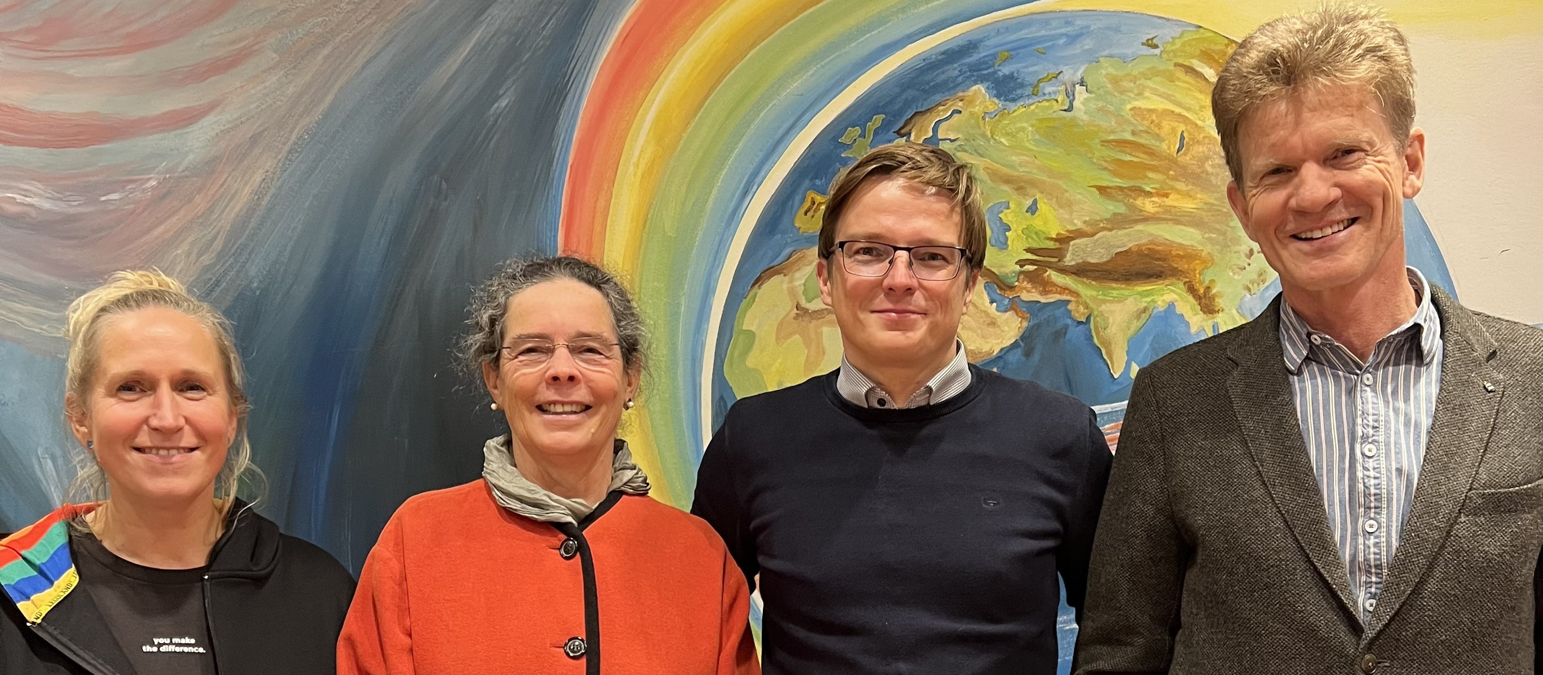 KI generiert: Das Bild zeigt vier lächelnde Personen, die vor einer farbenfrohen Wand mit einer Darstellung der Erde und einem Regenbogen stehen. Der Hauptinhalt des Bildes ist die Gruppe von Menschen, die gemeinsam posieren.