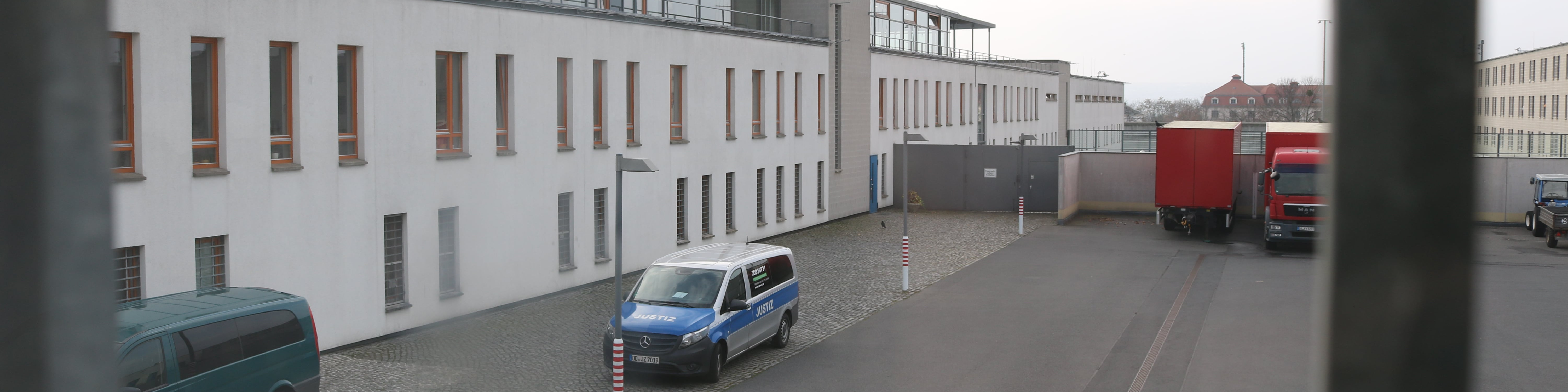 KI generiert: Das Bild zeigt einen Innenhof mit gepflastertem Boden, umgeben von Gebäuden mit weißen Fassaden und mehreren Fenstern. Im Vordergrund steht ein blauer Polizeiwagen, und im Hintergrund sind ein roter LKW sowie weitere Fahrzeuge zu sehen.