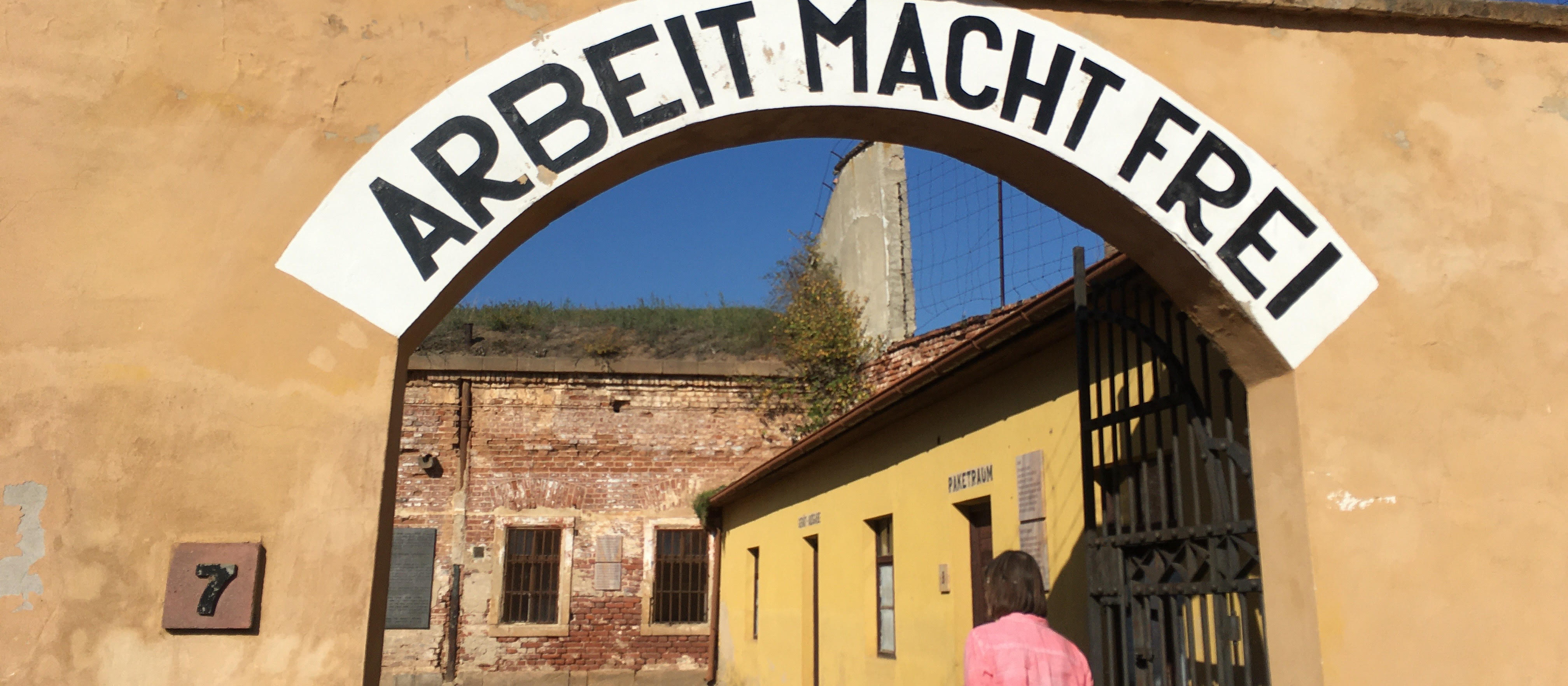 KI generiert: Das Bild zeigt den Eingang zum ehemaligen Konzentrationslager Dachau mit dem zynischen Spruch "Arbeit macht frei" über dem Tor. Im Hintergrund sind Lagergebäude und ein Wachturm zu sehen.
