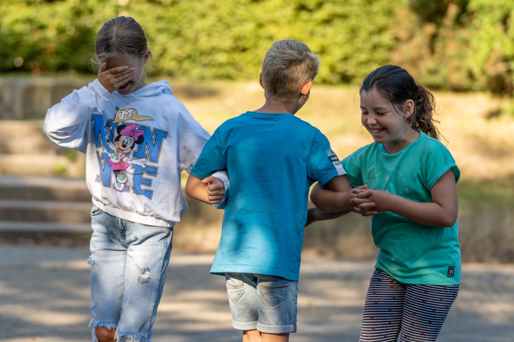 KI generiert: Das Bild zeigt drei Kinder, die draußen auf einem Weg spielen. Ein Mädchen mit einem Minnie-Mouse-Shirt bedeckt ihre Augen, während die anderen beiden Kinder sich an den Händen halten und lachen.