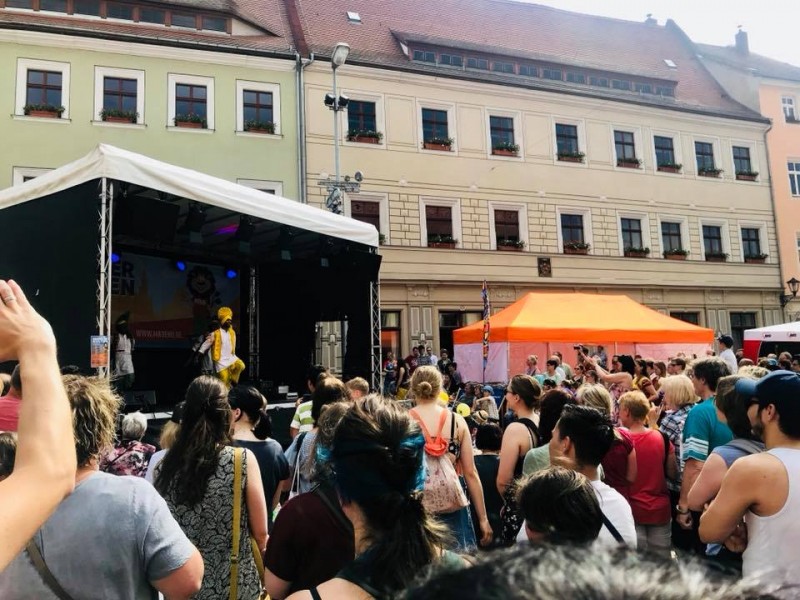 KI generiert: Das Bild zeigt eine Menschenmenge bei einem Open-Air-Konzert oder einer öffentlichen Veranstaltung in einer Stadt. Im Vordergrund ist eine Bühne mit einem Künstler zu sehen, während im Hintergrund historische Gebäude und ein weiteres Zelt zu erkennen sind.