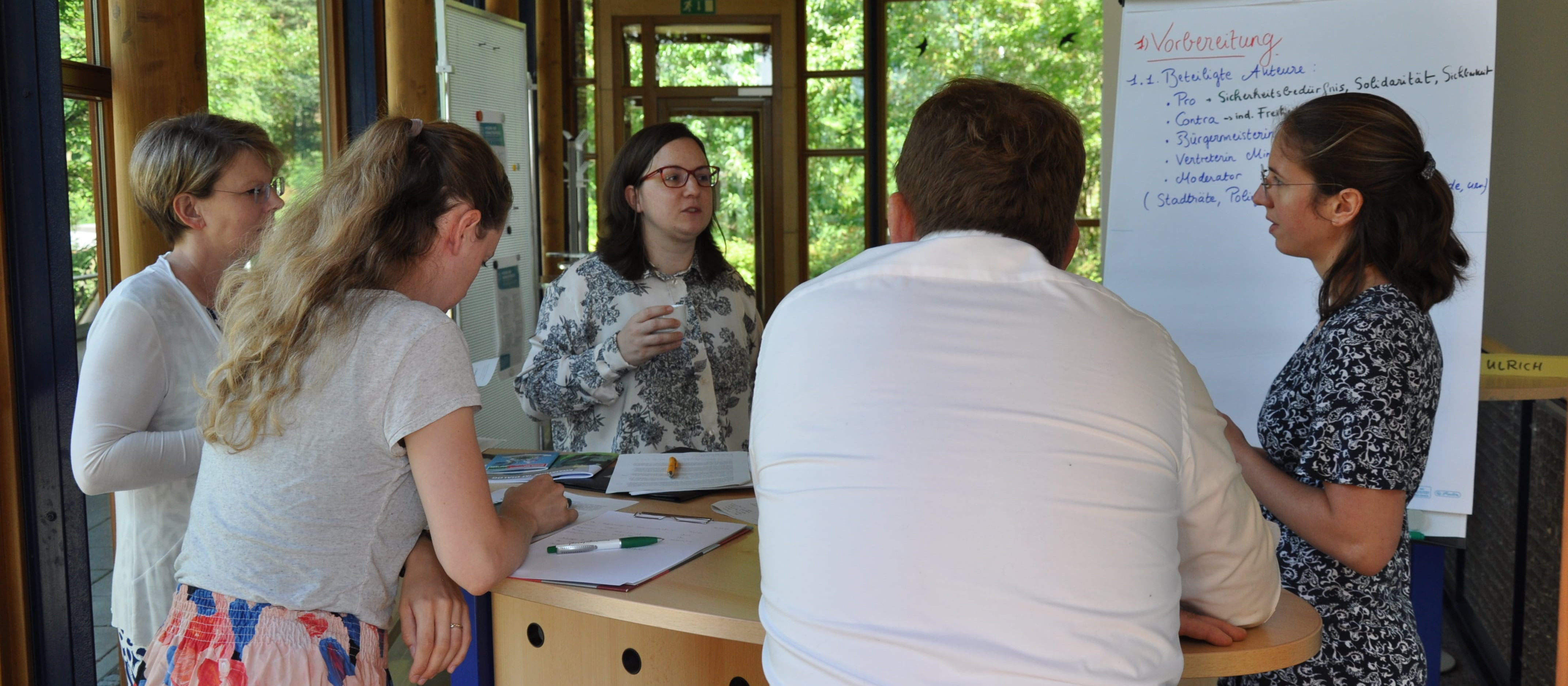 KI generiert: Das Bild zeigt eine Gruppe von Personen, die in einem hellen Raum mit großen Fenstern und Blick auf Grünflächen eine Besprechung oder ein Brainstorming abhalten. Im Vordergrund steht eine Person vor einem Whiteboard mit Notizen, während die anderen Teilnehmer aufmerksam zuhören oder sich Notizen machen.