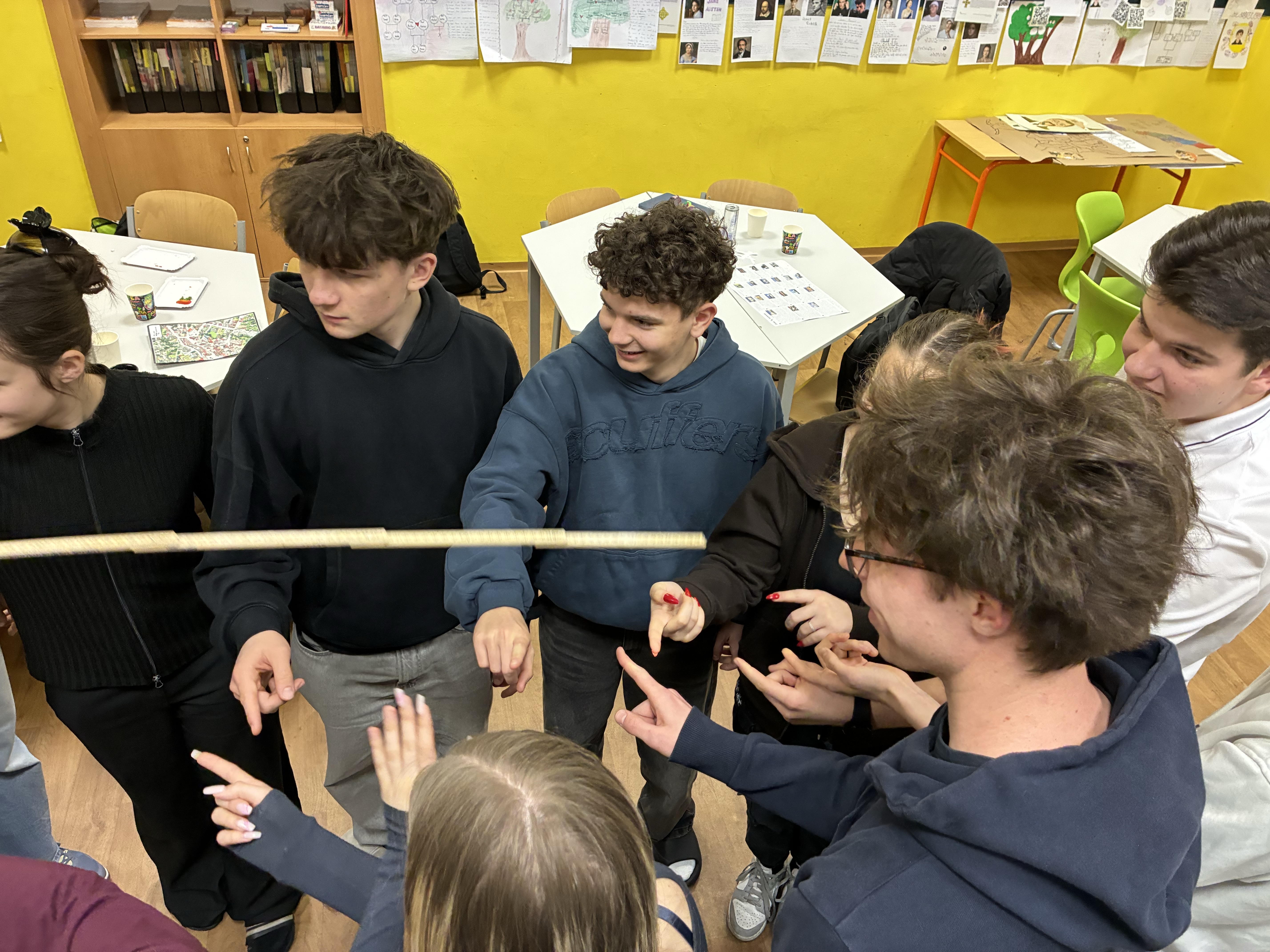 KI generiert: Das Bild zeigt eine Gruppe von Jugendlichen, die in einem Klassenzimmer zusammenstehen und sich auf eine Aktivität konzentrieren, bei der sie mit den Händen etwas zu halten oder zu balancieren scheinen. Der Hauptinhalt des Bildes ist die Interaktion und Zusammenarbeit der Gruppe während einer scheinbar spielerischen oder pädagogischen Übung.
