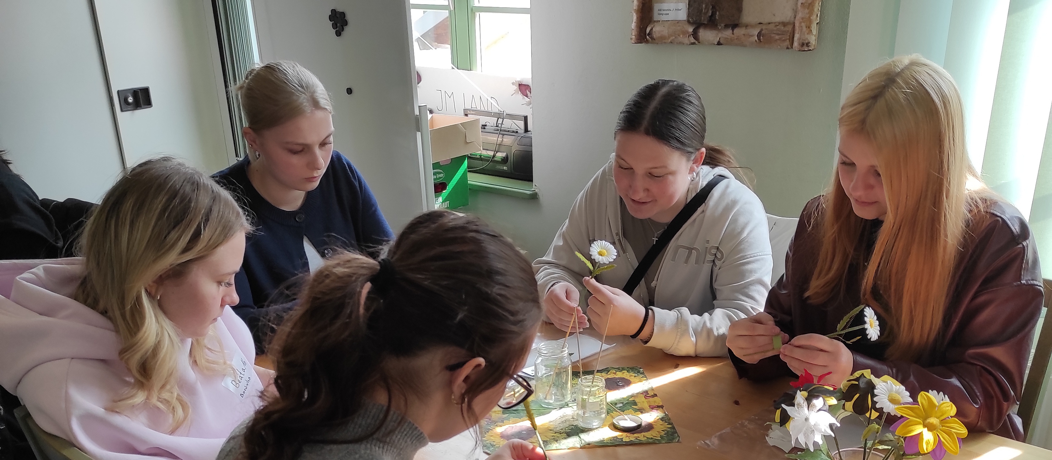 KI generiert: Das Bild zeigt eine Gruppe von fünf Personen, die an einem Tisch sitzen und gemeinsam Blumen oder Pflanzen in Gläsern arrangieren. Der Hauptinhalt des Bildes ist eine kreative Bastelaktivität in einer gemütlichen, gut beleuchteten Umgebung.