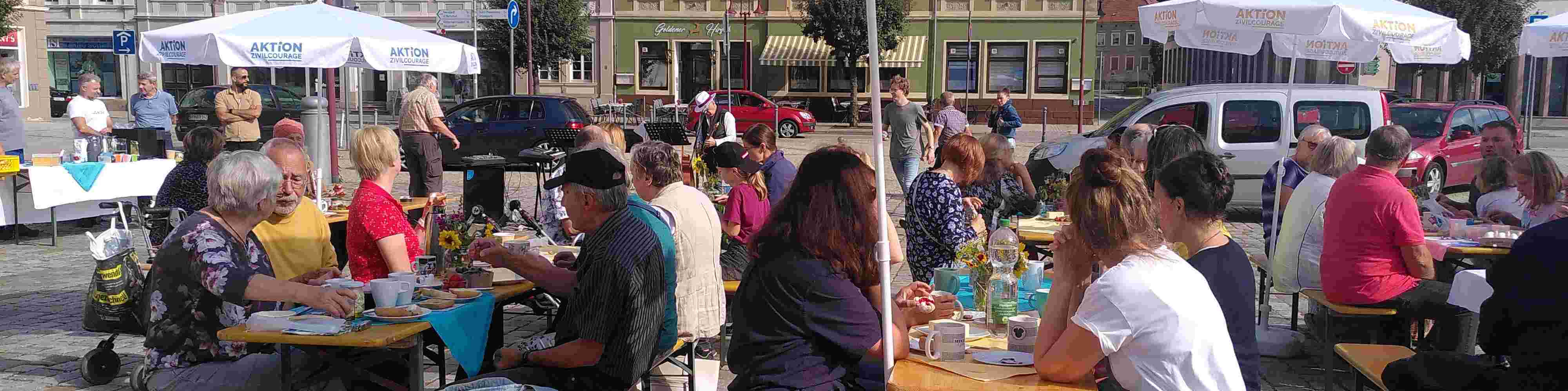 KI generiert: Das Bild zeigt eine belebte Außenbereichs-Szene mit vielen Menschen, die an langen Tischen sitzen und sich unterhalten, während sie Speisen und Getränke zu sich nehmen. Im Hintergrund sind bunte Gebäude und parkende Autos zu sehen, was auf ein geselliges Event oder eine öffentliche Veranstaltung in einer Stadt hindeutet.