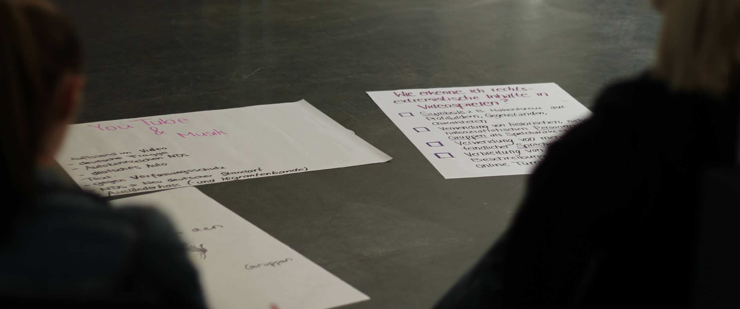 KI generiert: Das Bild zeigt zwei Personen, die an einem Tisch sitzen und auf mehrere Blätter Papier schauen, die auf dem Tisch liegen. Die Blätter enthalten handschriftliche Notizen und Texte, wobei eines der Blätter den Titel "Tube & Mask" trägt.