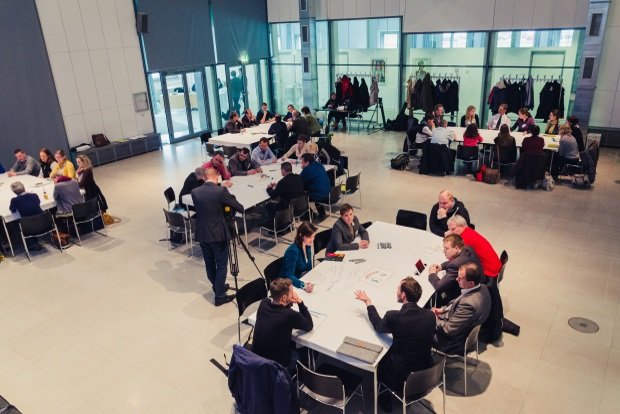 KI generiert: Das Bild zeigt eine Gruppe von Menschen, die in kleinen Gruppen an mehreren runden Tischen in einem hellen, modernen Raum sitzen und sich unterhalten. Der Hauptinhalt des Bildes ist eine Versammlung oder ein Workshop, bei dem die Teilnehmer in Diskussionen oder Arbeitsgruppen engagiert sind.