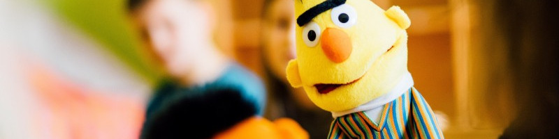 KI generiert: Das Bild zeigt eine Nahaufnahme einer Puppe, die Bert aus der Sesamstraße darstellt. Im Hintergrund sind unscharf zwei Personen zu erkennen.