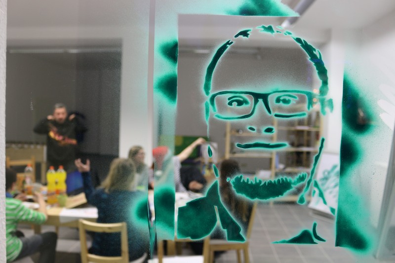 KI generiert: Das Bild zeigt ein Klassenzimmer oder einen Workshop, in dem eine Gruppe von Menschen an Tischen sitzt und arbeitet. Im Vordergrund ist ein Glas oder eine durchsichtige Oberfläche mit einem großen, grünen Graffiti-Porträt eines Mannes mit Brille zu sehen.