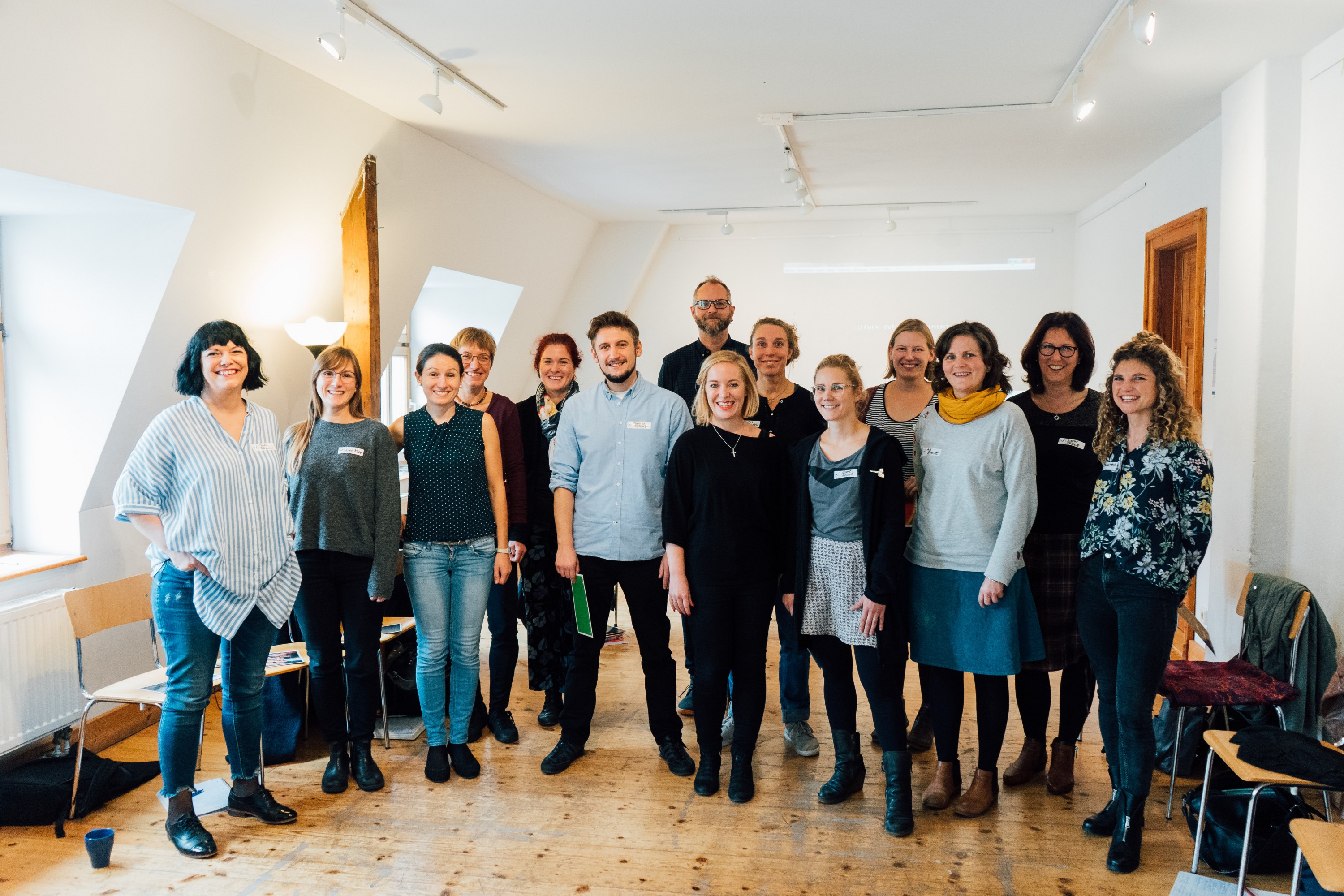 KI generiert: Das Bild zeigt eine Gruppe von Personen, die in einem hellen Raum mit Holzboden und schrägen Wänden stehen und für ein Gruppenfoto posieren. Der Hauptinhalt des Bildes ist die Gruppe von Menschen, die gemeinsam ein Foto machen.