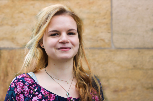 KI generiert: Das Bild zeigt eine lächelnde junge Frau mit langen blonden Haaren, die vor einer steinernen Wand steht. Sie trägt ein buntes, blumiges Oberteil und einen Halsketten-Schmuck.