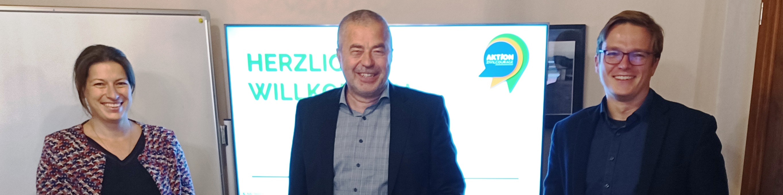 KI generiert: Das Bild zeigt drei lächelnde Personen, die vor einem Plakat mit der Aufschrift "HERZLICH WILLKOMMEN" und dem Logo "AKTION" stehen. Der Hauptinhalt des Bildes ist eine Begrüßungsszene, möglicherweise bei einer Veranstaltung oder einem Meeting.
