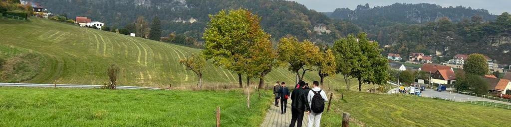 KI generiert: Das Bild zeigt eine Gruppe von Personen, die auf einem Feldweg durch eine ländliche, grüne Landschaft mit sanften Hügeln und verstreuten Bäumen spazieren. Im Hintergrund sind einige Häuser und ein Wald zu sehen.