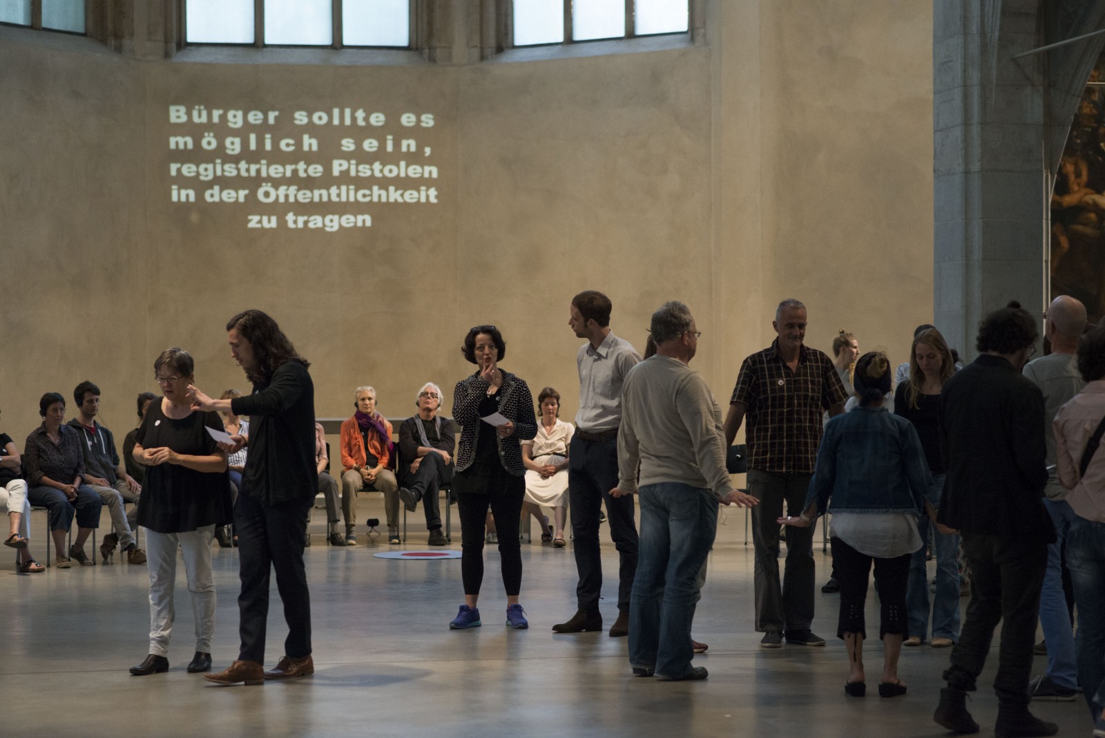 KI generiert: Das Bild zeigt eine Gruppe von Menschen in einer großen, hallenartigen Umgebung, die sich zu unterhalten und zu diskutieren scheinen. Im Hintergrund ist eine Projektion mit einem deutschen Text, der zur Registrierung von Pistolen in der Öffentlichkeit auffordert.
